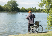 Image Of A Homeless Man Standing Next To His Wheel Chair By The Lake With A Fishing Rod In The Water On A Warm Summer Morning,