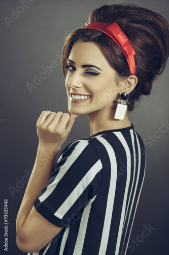 Naklejka na meble Beautiful persuasive lady tempting with retro sixties fashion style. Enticing smiling young woman in striped attire, red headband and retro updo hairstyle, winking at camera over dark background.