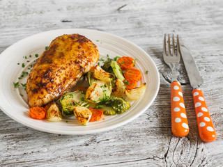 baked chicken breast with brussels sprouts, onions and carrots on a white plate on wooden surface. Healthy food