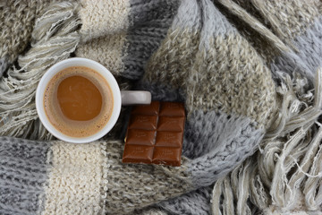 
Hot chocolate in coffee mug cup with a grey, ivory, ecru knitted scarf blanked wrapped around it and chocolate bar.