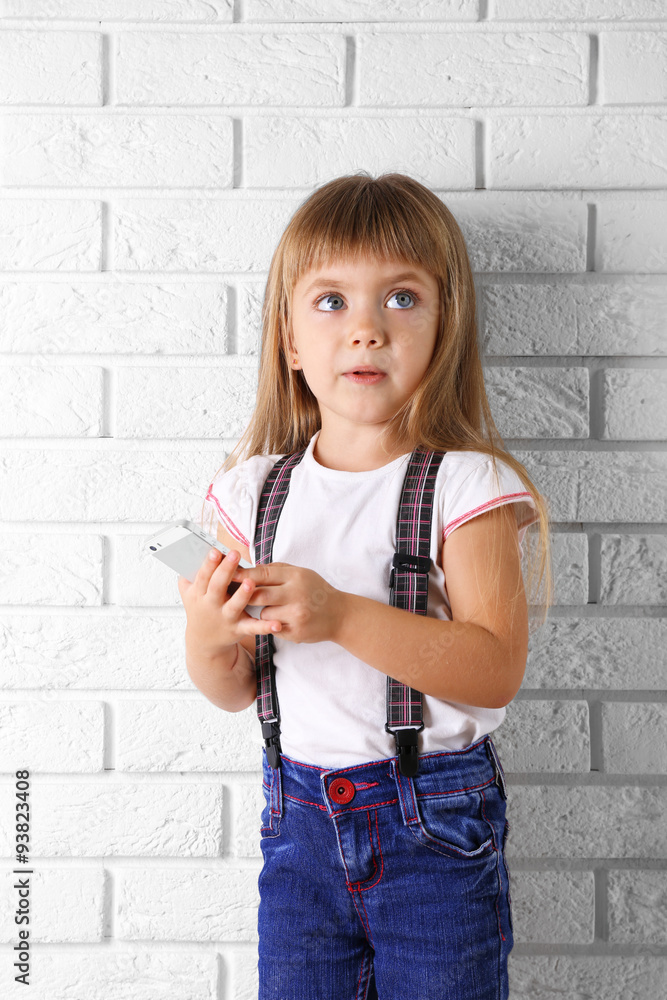 Beautiful little girl with mobile phone on light wall background