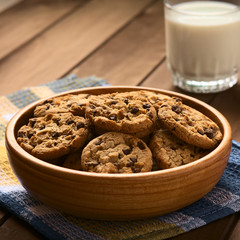 Wall Mural - Chocolate chip cookies in wooden bowl with a glass of cold milk in the back, photographed on cloth on wood with natural light (Selective Focus, Focus in the middle of the cookies)