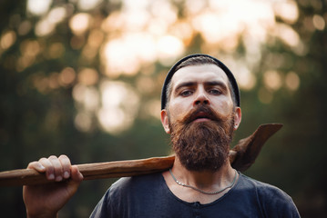 Wall Mural - bearded brutal man in warm hat lumberjack with an ax in a forest at sunset, and the thick forest in the rain, closeup portrait