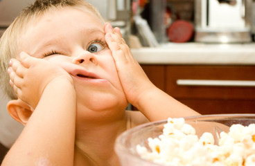 Funny baby bow with .skinned elbow and one wide opened eye on a kitchen background near the bowl of popcorn is making faces