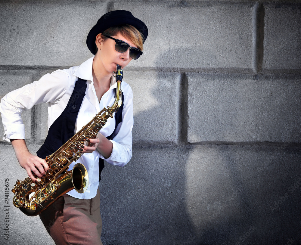 Attractive woman in hat plays saxophone on brick wall background