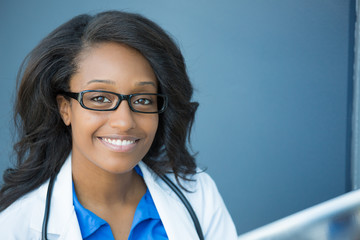 Closeup headshot portrait of friendly, smiling confident female healthcare professional with lab coat, glasses, and stethoscope. Isolated hospital clinic background. Time for an office visit