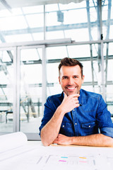 young handsome smiling architect looking at camera sitting at desk over some blueprints