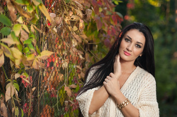 Beautiful woman in white posing in autumnal park. Young brunette woman spending time in autumn near a tree in forest. Long dark hair attractive woman smiling with faded leaves around her