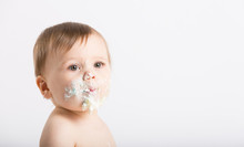 A Cute 1 Year Old Sits In A White Studio Setting. Close Up Of A Baby With A Face Full Of Cake And Frosting. He Is Only Dressed In A White Diaper