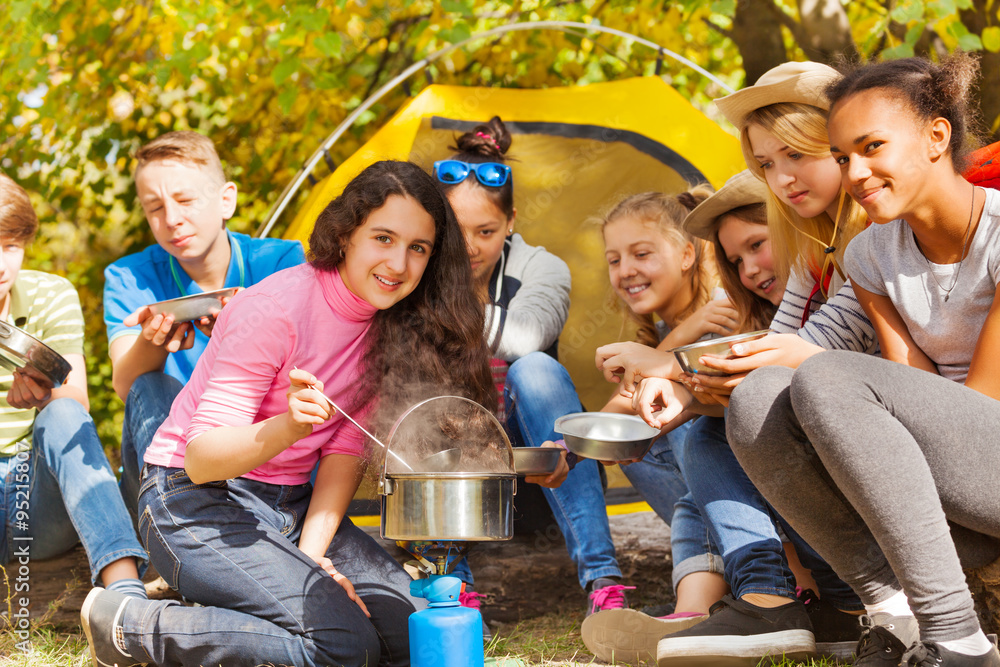 Teenagers cook soup in metallic pot during camping - obrazy, fototapety, plakaty 