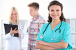 © rogerphoto - Beautiful young smiling female doctor standing at hospital with doctor and patient on the background. Medical concept.