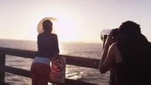 Black Woman Looking Through Telescope At Pier While Friend Leans Against The Railing And Looks At The Ocean Water