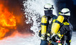Feuerwehr - Feuerwehrmänner löschen ein großes Feuer, sie stehen vor einer Feuerwand und tragen Schutzkleidung