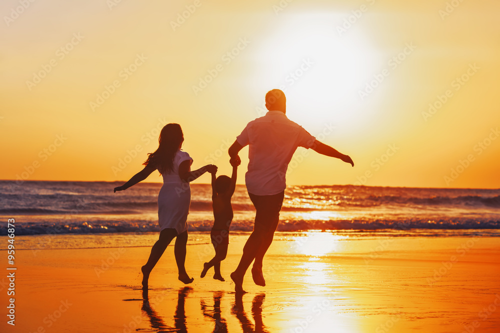 Happy family - father, mother, baby son hold hands and run with fun along edge of sunset sea on black sand beach. Active parents and people outdoor activity on tropical summer vacations with children. - obrazy, fototapety, plakaty 