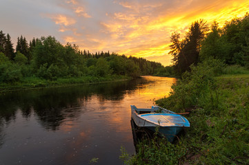 Bright dramatic sunset over river with forest along riverside and boat in foreground at white night. Arkhangelsky region, Russia.
