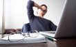 man with his hand holding his face taking a brake from working with laptop computer and notebook with eye glasses on wooden desk. concept of stress/rest/tension/failed/discourage