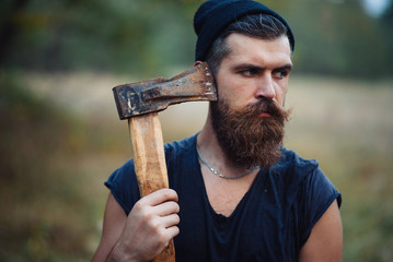 Canvas Print - Brutal brunette bearded man in warm hat with a hatchet in the woods on a background of trees