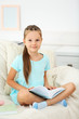 © Africa Studio - Beautiful little girl sitting on sofa with book, on home interior background