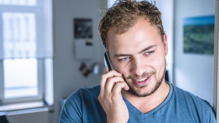 Wall Mural - Young man making a phone call from his office. Handsome young man talking on the phone smiling happy wearing leisure outfit. Urban male in his 20s. 