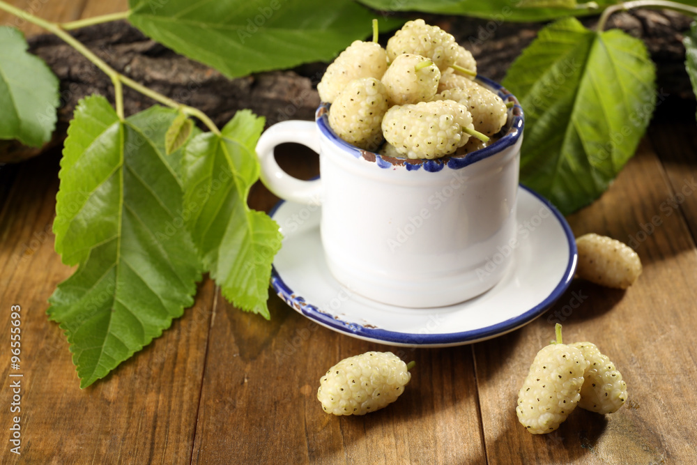 Ripe mulberries in mug with green leaves on table close up