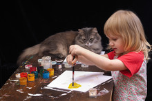 Ребенок и кот рисуют. Маленький ребенок со светлыми волосами рисует красками на столе, на белой бумаге. На столе сидит серый кот и внимательно наблюдает, как рисует  Девочка  солнце желтой краской