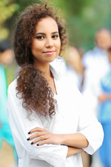 Poster - Attractive young woman doctor with clipboard in hands against group of medics, outdoors