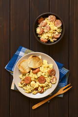 Wall Mural - Overhead shot of scrambled eggs made with chorizo slices and onion on plate with toasted baguette slices, wooden fork on the side, photographed on dark wood with natural light