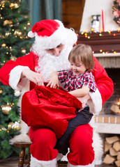 Wall Mural - Santa Claus pulls a present out sack to child near the fireplace and Christmas tree at home