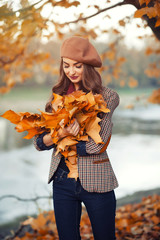 slender young brunette beautiful girl with long hair in a beret with a scarf and coat walks in autumn park with a lot of yellow and orange leaves