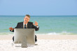 © Michael O'Keene - A business man in a suit is sitting on the beach with a big computer in front of him and he has a very happy look on his face