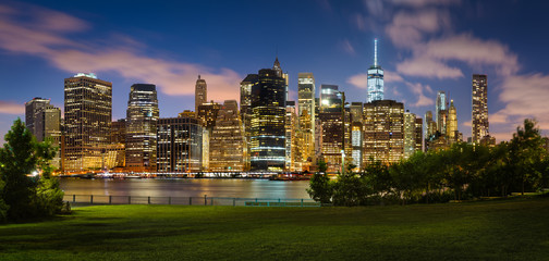 Fototapete - Lower Manhattan skyline and green lawn of Brooklyn Bridge Park at twilight.Illuminated skyscrapers of the Financial District reflect on the East River. New York City.