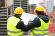 © cineberg - Back turned construction workers with yellow hardhat and safety jacket checking blueprint