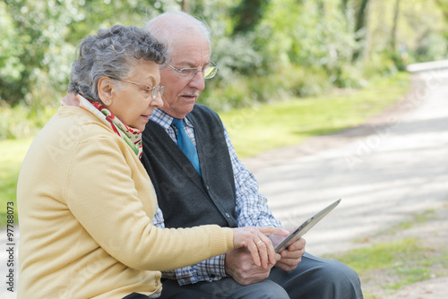 Fotografie, Obraz Elderly couple sat on bench looking at tablet