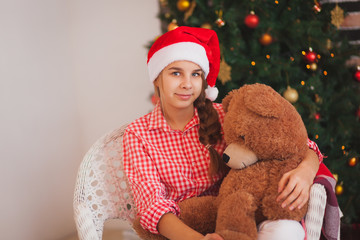  holidays, presents, christmas, childhood and people concept - smiling girl in santa helper hat with teddy bear over lights background