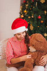  holidays, presents, christmas, childhood and people concept - smiling girl in santa helper hat with teddy bear over lights background