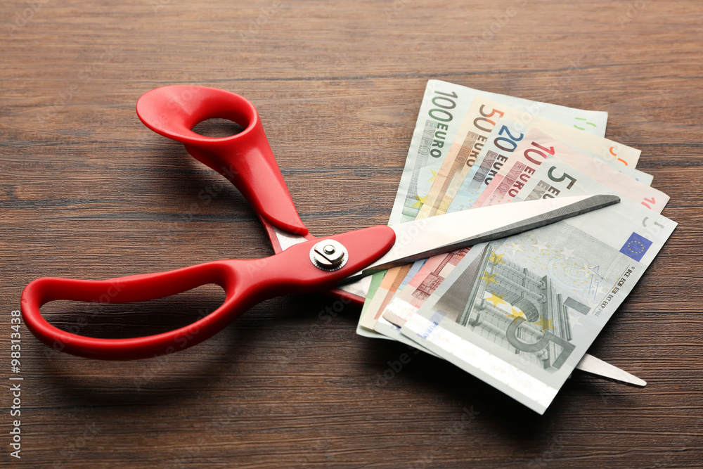 Red scissors cut money on wooden background. Financial concept