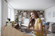 © Westend61 - Smiling woman at home with cell phone, digital tablet and laptop on secretary desk