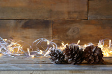 Pine Cones Next To Gold Garland Lights On Wooden Background. Copy Space
