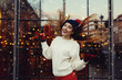 Street fashion portrait of smiling beautiful young woman playing with her long hair. Lady wearing classic winter knitted clothes. Festive  garland lights. Magic snowfall effect. Toned