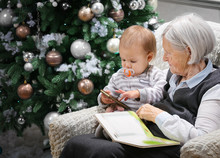 Senior Woman Reading A Book To Her Baby Grandson Beside A Christmas Tree, The Boy Is Playing With A Smartphone