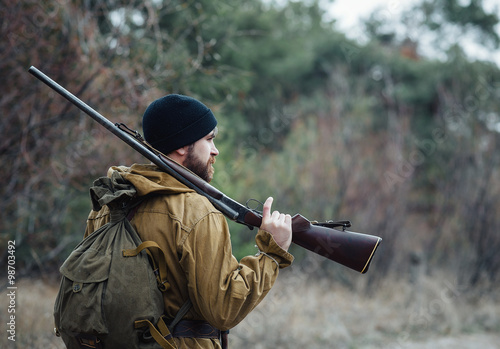 Fotografie, Obraz brutal hunter, bearded man in warm hat with a gun in his hand, a knife a backpac