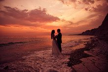 A Just Married Couple Staying At The Beach. Romantic Wedding Sunset. Splashing Ocean Waves And Beautiful Sky