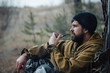 © endrews21 - brutal hunter, bearded man in warm hat with a gun in his hand, a knife a backpack and smoking pipe in the wild forest in the autumn