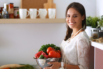 Wall Mural - Smiling young woman holding vegetables standing in kitchen