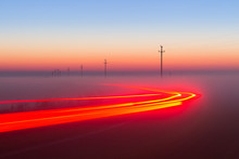 Long Exposure Red Car Light Trails On A Road Outside At Foggy Night On Blue Hour With Electrical Power Lines And Pylons Disappear Over The Horizon