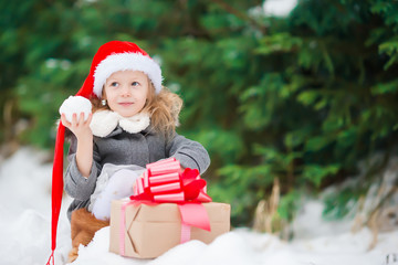 Wall Mural - Happy little girl in santa hat with christmas box gift in winter day outdoors on Xmas eve