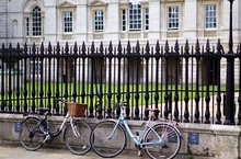 Two Women's Town Bicycles, Facing Each Other, Leaning Against An Ornamental Iron Railing In Front Of A Historic University Building In Cambridge, England