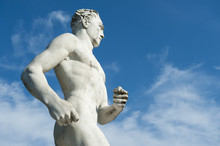 Ancient Marble Statue Of Muscular Athlete Running Against Bright Blue Sky