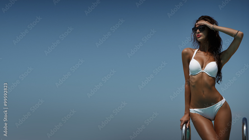 Elegant woman in the white bikini on the sun-tanned slim body is posing near the swimming pool in a good hot weather - obrazy, fototapety, plakaty 
