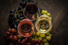 Three Wineglasses Of Red, Rose And White Wine With Three Kinds Of Grapes On Brown Wood Textured Table